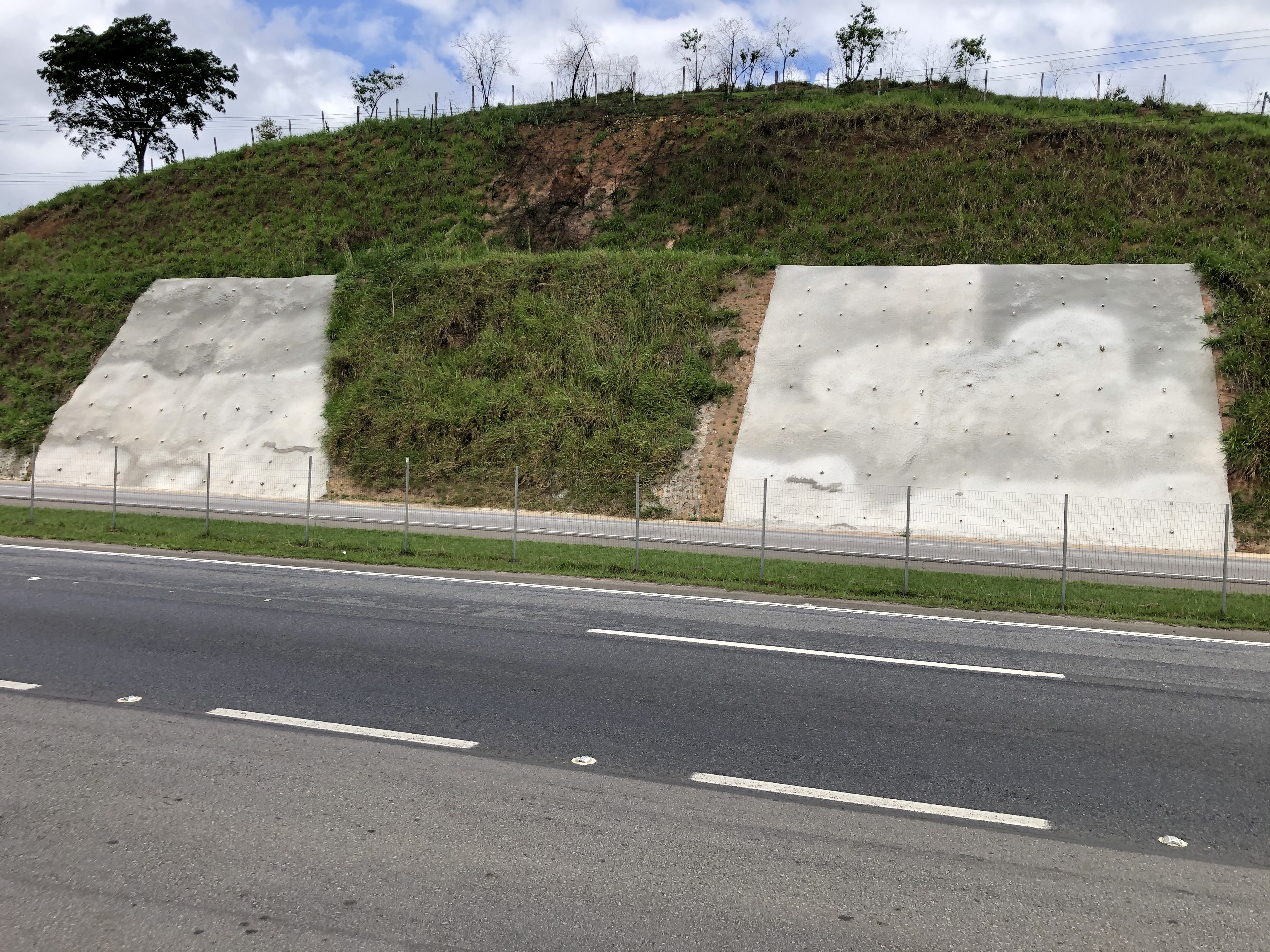 Muros de contenção de taludes em concreto construídos pela Novacap para estabilização de encosta ao lado de rodovia, vegetação no talude e estrada em primeiro plano.