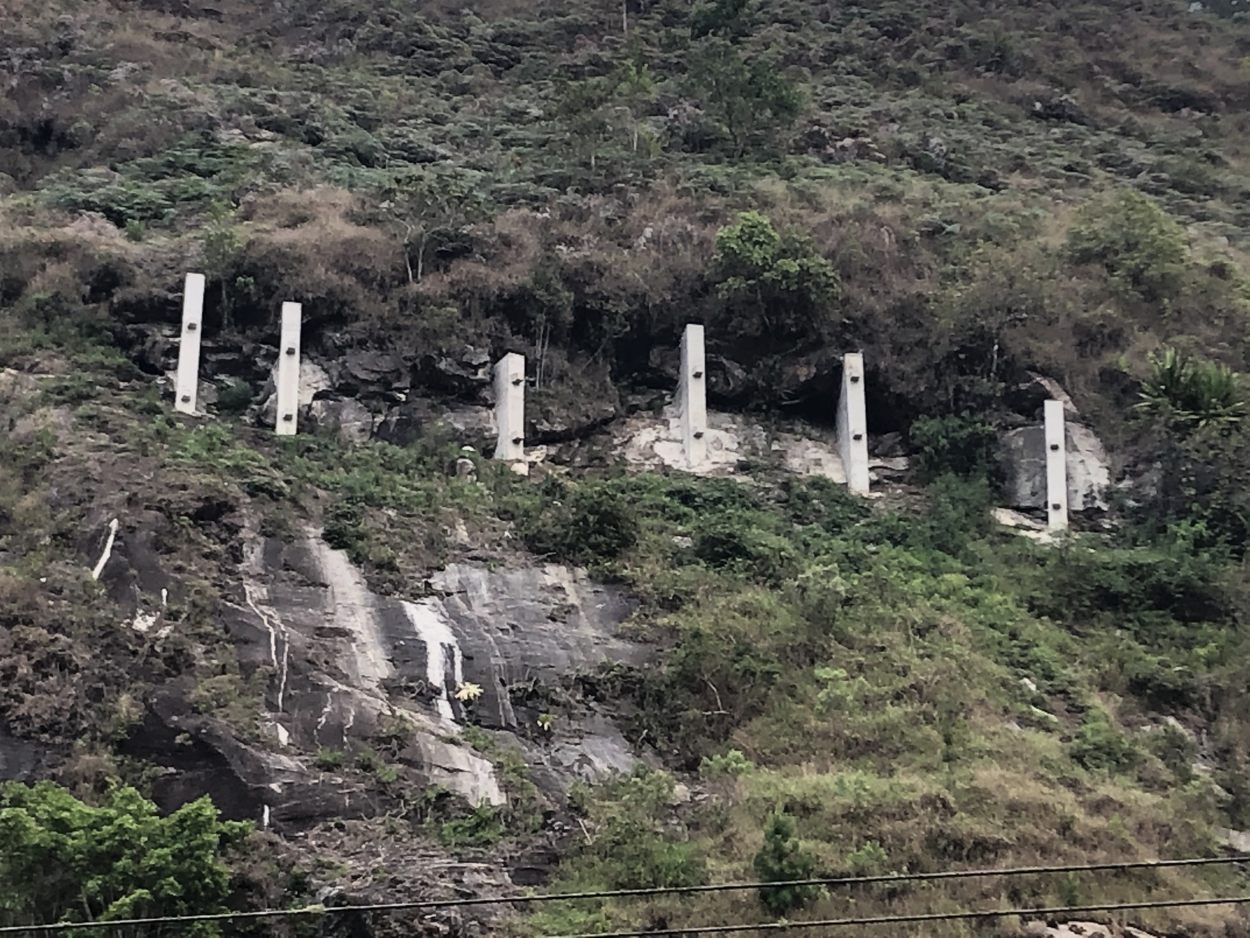 Muros de gabião sendo construídos pela Novacap para contenção de talude rochoso ao lado de rodovia.  Talude parcialmente coberto por lona e terra, cones de sinalização na via e vegetação na encosta.