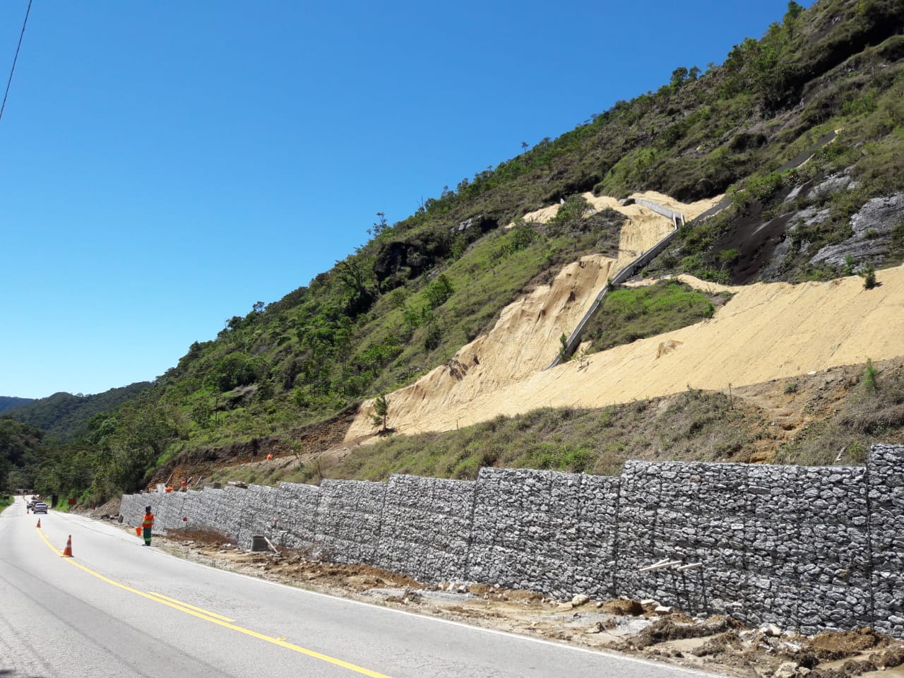 Pilares de concreto armado sendo instalados pela Novacap em talude rochoso para obra de estabilização. Colunas verticais fixadas na rocha com vegetação e encosta rochosa ao fundo